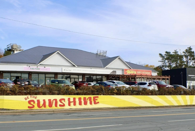 A large outdoor mural painted on a retaining wall in front of a building. The background is a bright, golden yellow with bold rays extending outward like sunshine. Centered in the mural, the words "Be the Sunshine" are painted in a vivid red, standing out against the warm backdrop. The design is simple yet striking, evoking warmth, positivity, and encouragement for passersby.