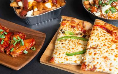 Pizza and wings served on a wooden cutting board sitting on a table. There is also poutine in the background.